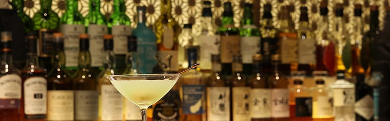 Image of cocktail glasses on a bar counter lined with many bottles of alcohol